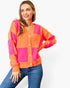 A woman wearing a bright orange and hot pink checkered knit cardigan with light-colored buttons, paired with blue jeans, against a plain white background.