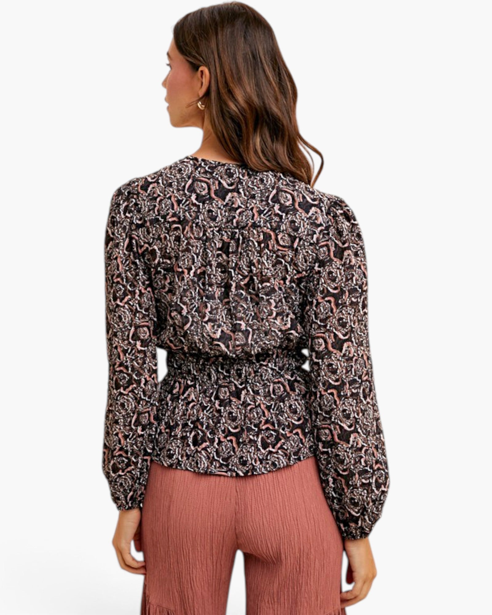 A woman shown from the back wearing a long-sleeve patterned blouse with a gathered waist and dusty rose pleated pants, against a plain light gray studio background.