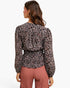A woman shown from the back wearing a long-sleeve patterned blouse with a gathered waist and dusty rose pleated pants, against a plain light gray studio background.