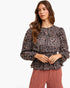 A woman modeling a long-sleeve blouse with a black, brown, and beige paisley pattern, paired with dusty rose trousers, against a plain white studio background.