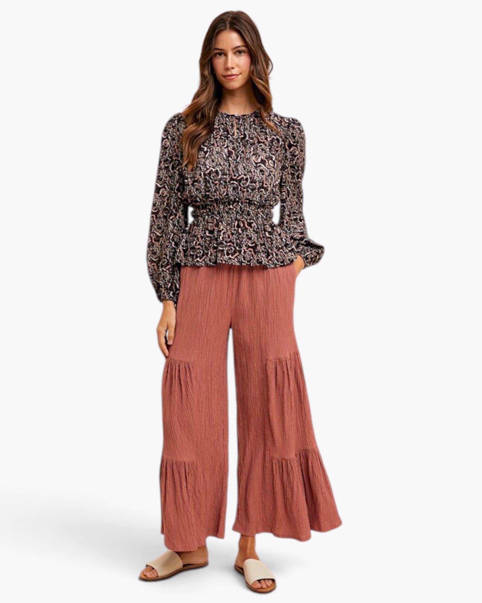 A woman modeling a long-sleeve black and beige patterned blouse paired with dusty rose wide-leg pleated pants and beige slide sandals, set against a plain white studio background.