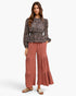 A woman modeling a long-sleeve black and beige patterned blouse paired with dusty rose wide-leg pleated pants and beige slide sandals, set against a plain white studio background.