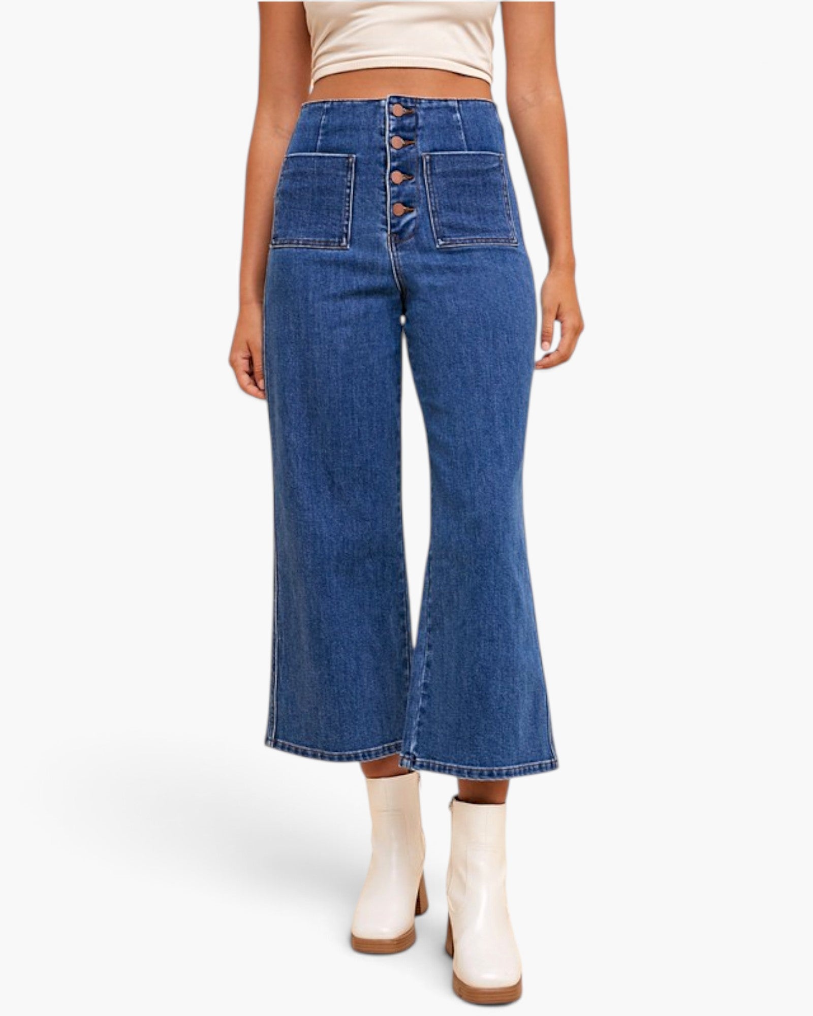 A model wearing high-waisted, wide-leg blue denim jeans with a button-fly front and front patch pockets, paired with cream ankle boots against a plain white studio background.