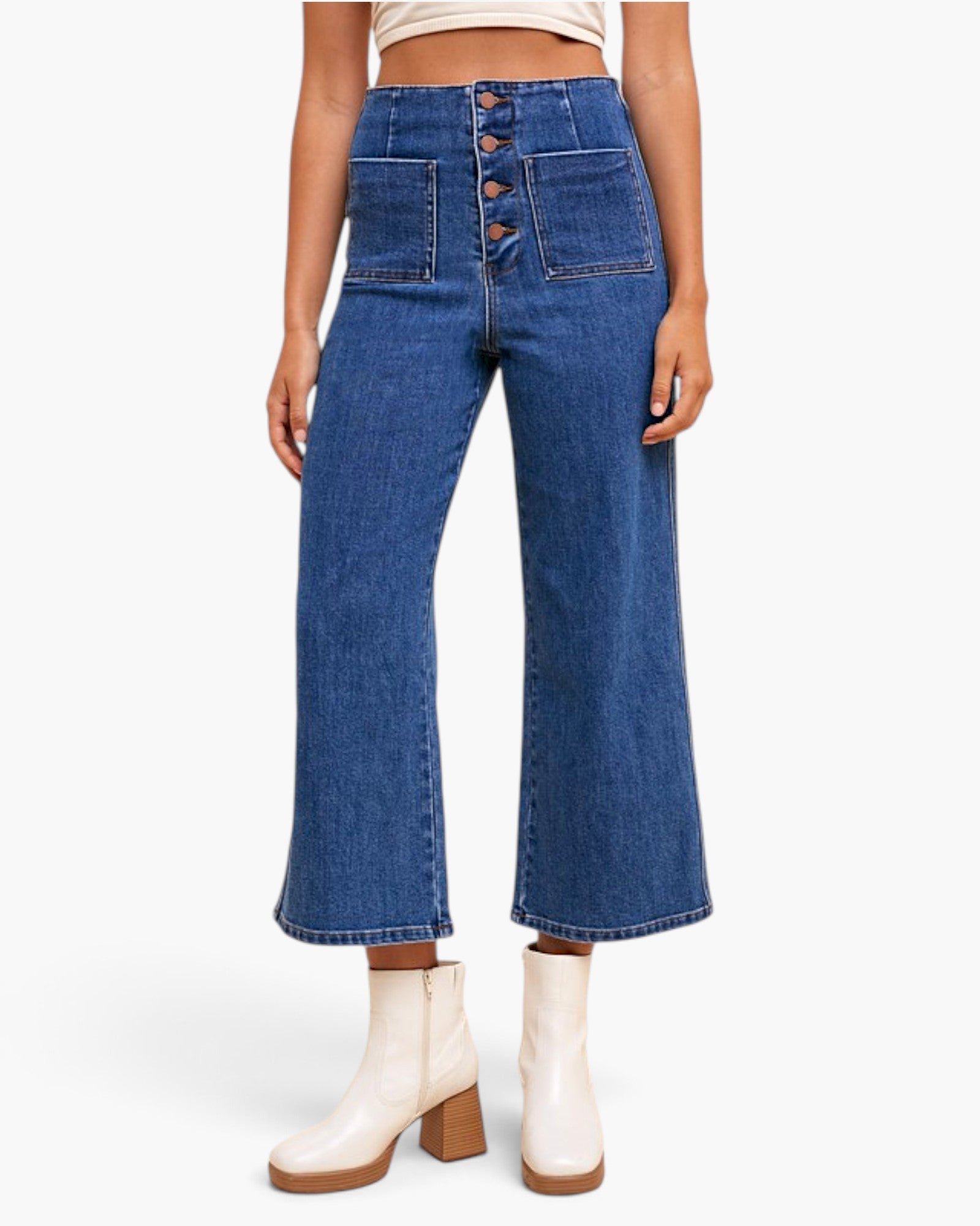 A model wearing a pair of high-waisted, wide-leg blue denim jeans with a button fly and front patch pockets, paired with white heeled ankle boots against a light gray studio background.