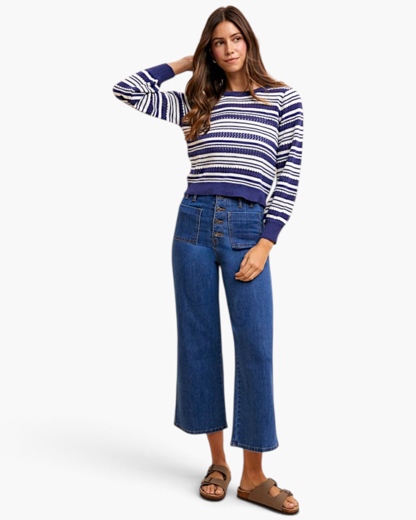 A woman modeling a navy and white horizontally striped long-sleeve knit sweater paired with blue cropped jeans and brown sandals, set against a plain light gray studio background.