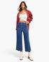 A woman modeling a pair of high-waisted, wide-leg blue jeans with a button fly, styled with a cream crop top, a red plaid cardigan, and white ankle boots against a plain white studio background.