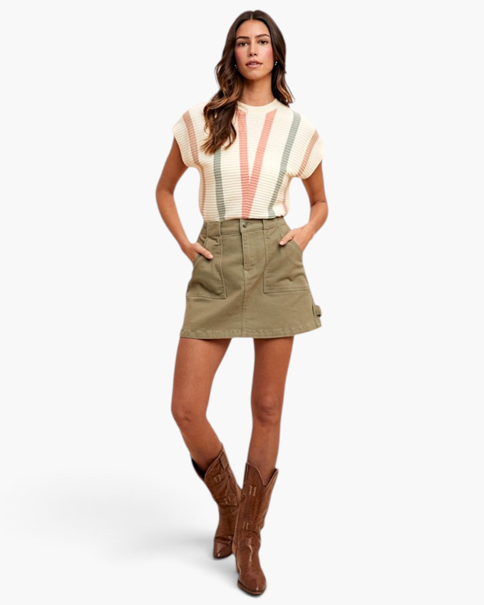 A woman modeling a short-sleeve knit top with cream, pink, and sage green stripes, paired with an olive green mini skirt and brown cowboy boots, against a light gray studio background.