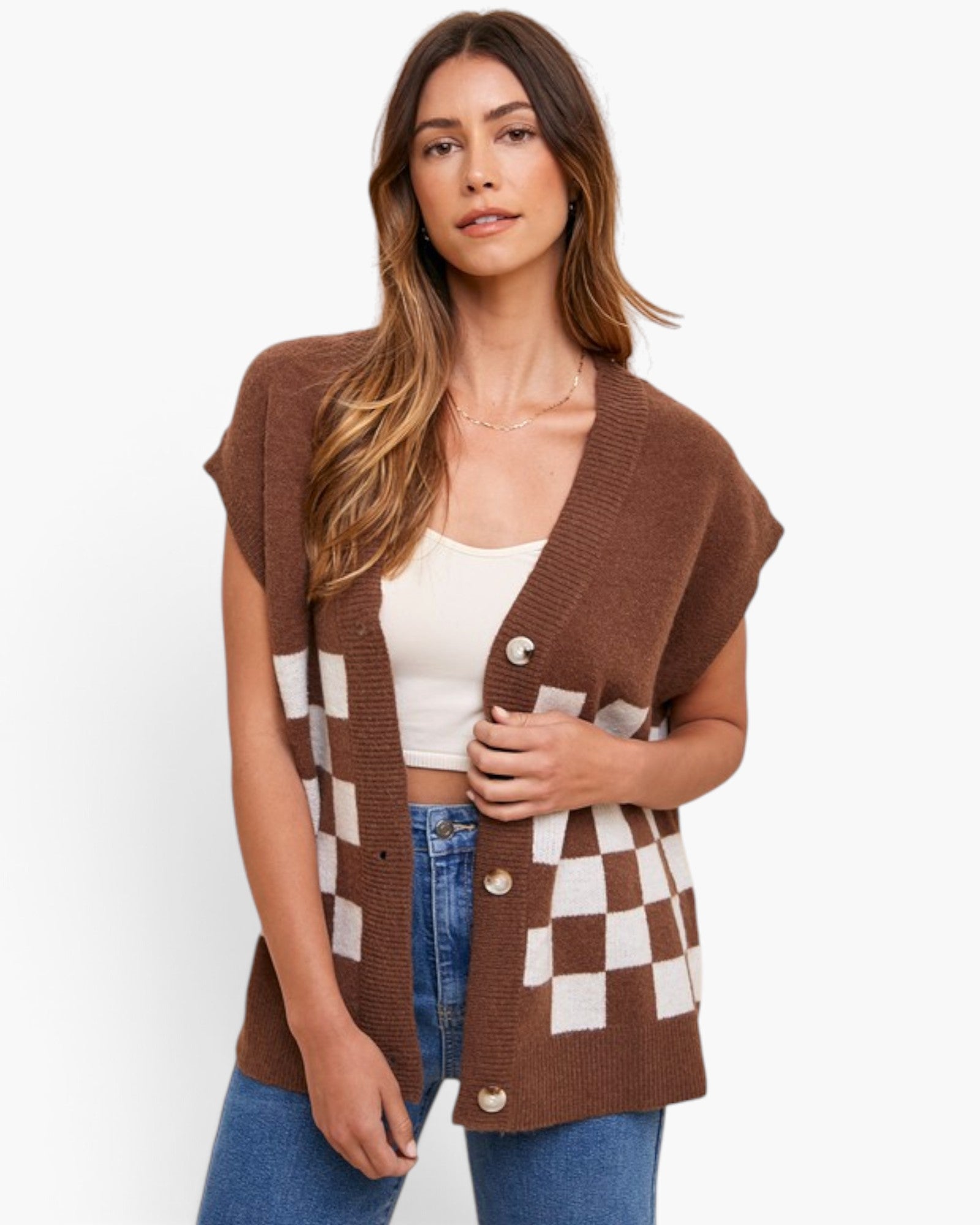 A woman modeling a brown sleeveless knit cardigan vest with white checkerboard pattern panels and front buttons, worn over a white crop top and blue jeans against a plain white background.