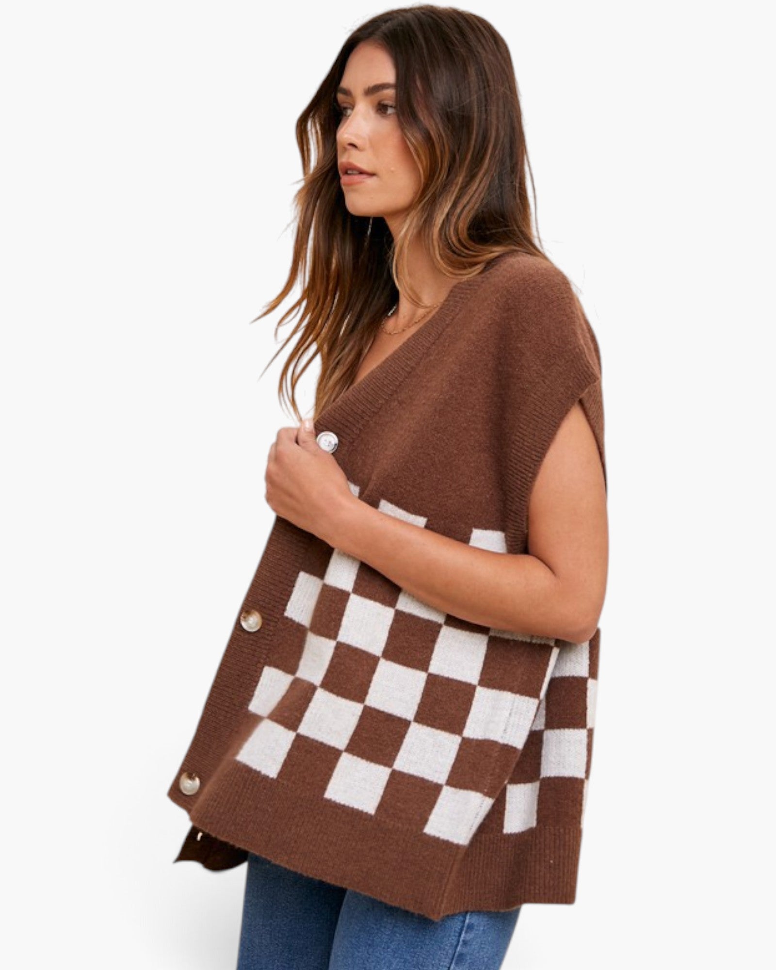 A woman wearing a brown knit poncho-style cardigan with a brown and white checkerboard pattern and gold-tone buttons, paired with blue jeans, against a plain white studio background.