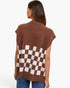 A woman viewed from the back wearing a brown knit poncho-style top with a large brown-and-white checkerboard pattern across the lower back, paired with blue jeans, against a plain light background.