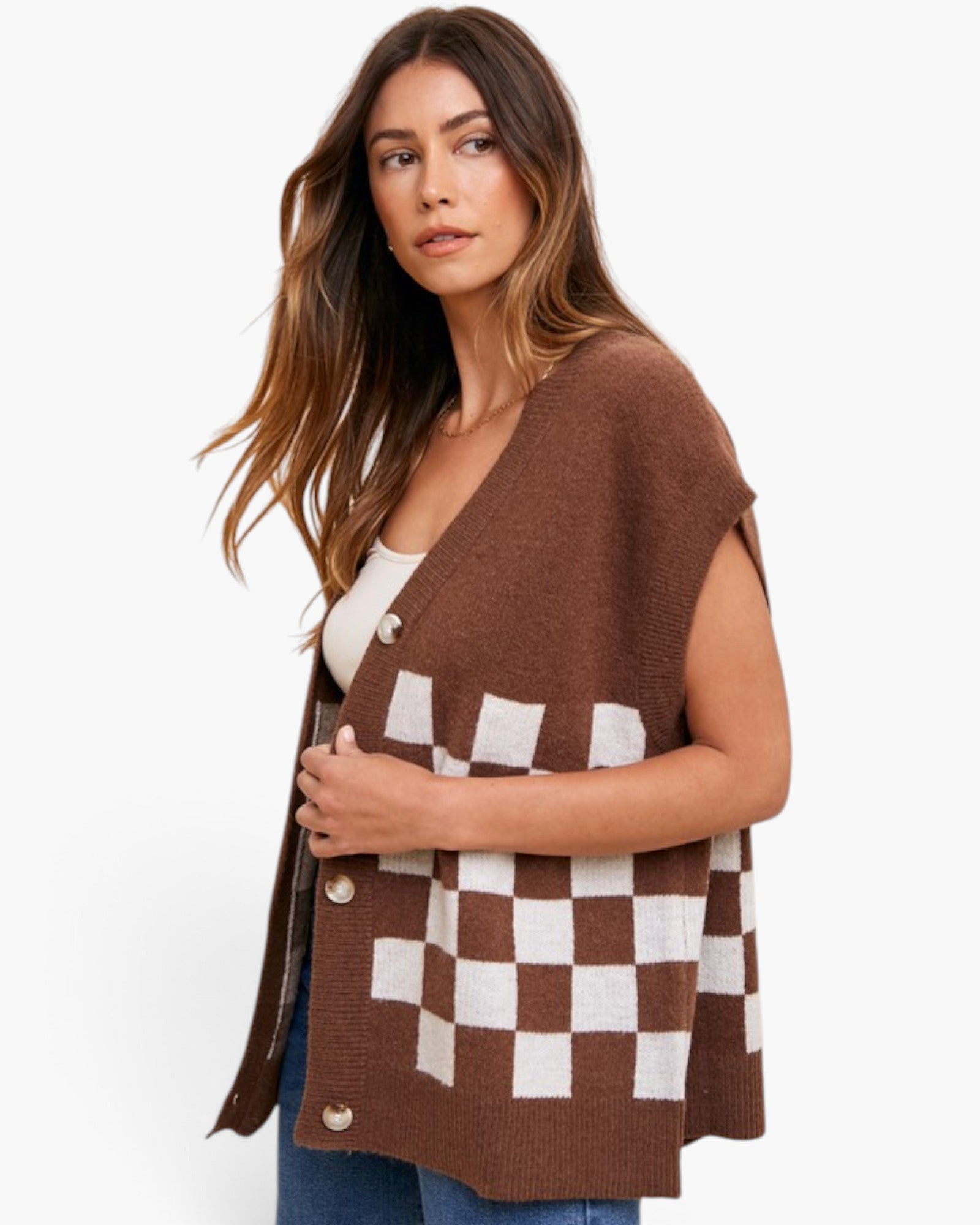 A woman wearing a brown knit cardigan vest with a white checkerboard pattern and silver buttons, styled over a white top and blue jeans against a plain white background.