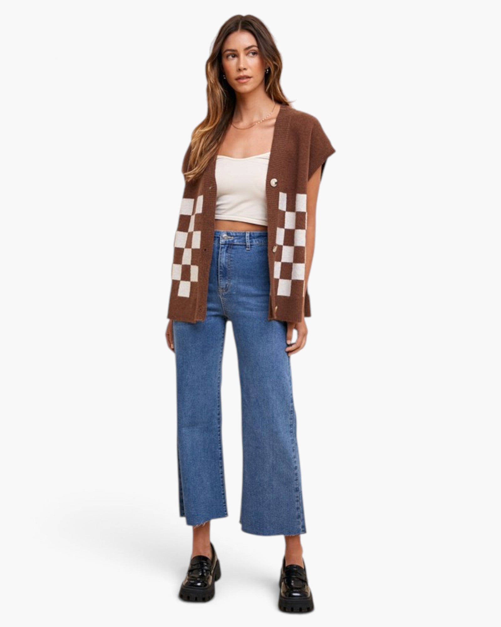 A woman stands against a light gray studio background wearing a brown sleeveless knit cardigan with beige-and-white checkerboard panels, a white cropped tank top, blue wide-leg cropped jeans, and black loafers.