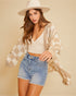Woman wearing a beige cardigan, white top, and denim shorts on a beige background