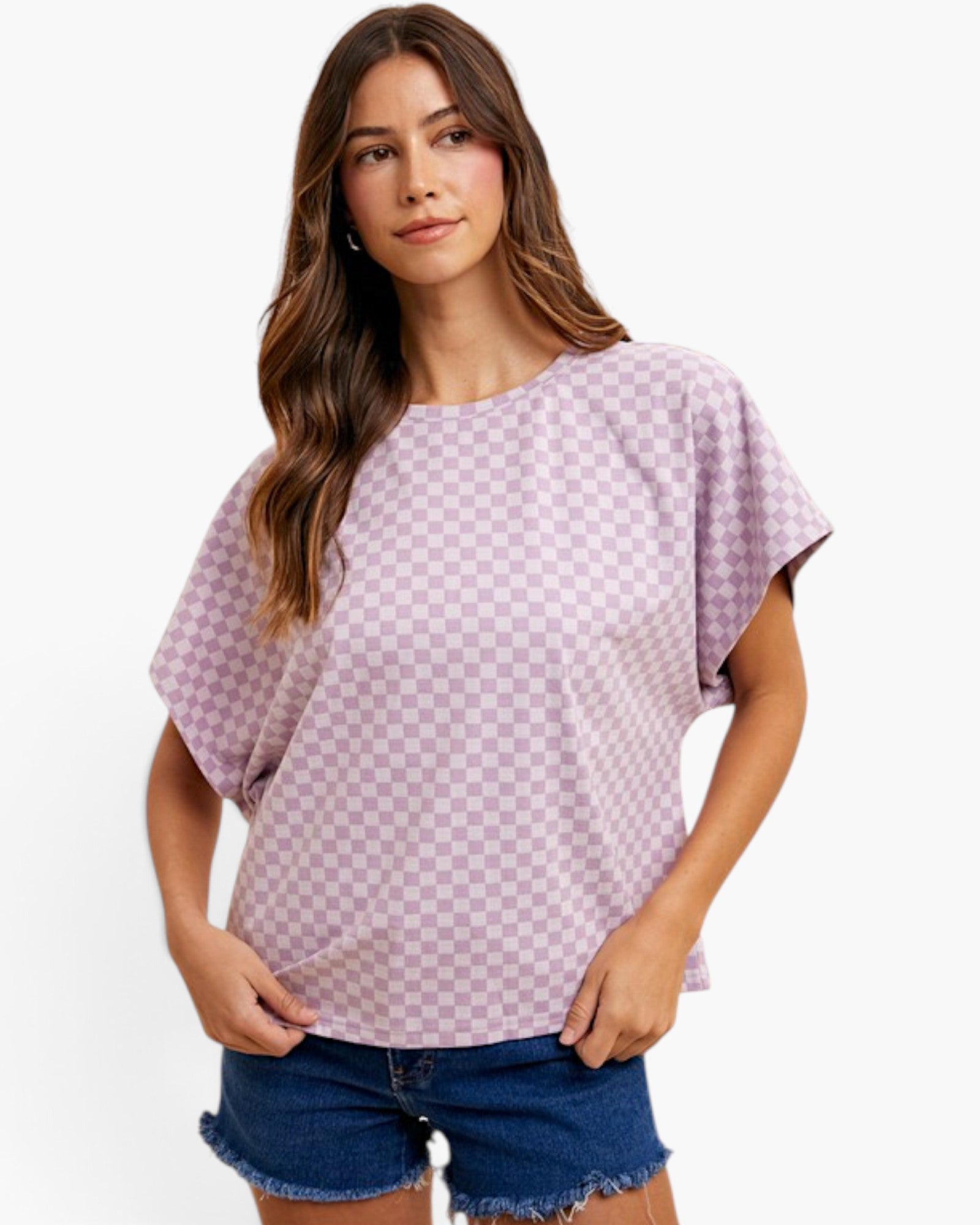 A woman modeling a lavender and light pink checkerboard patterned short-sleeve t-shirt with blue denim shorts against a plain white background.