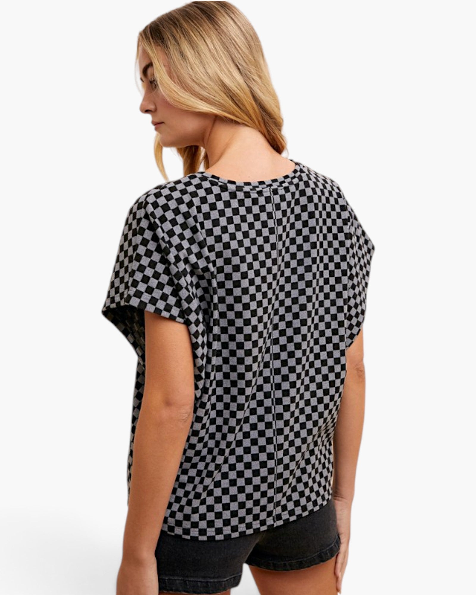 A woman shown from the back wearing a loose black and white checkerboard patterned top and dark denim shorts against a plain white studio background.