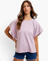 A woman modeling a lavender and white checkerboard short-sleeve top with a relaxed fit, paired with denim shorts, against a plain white background.