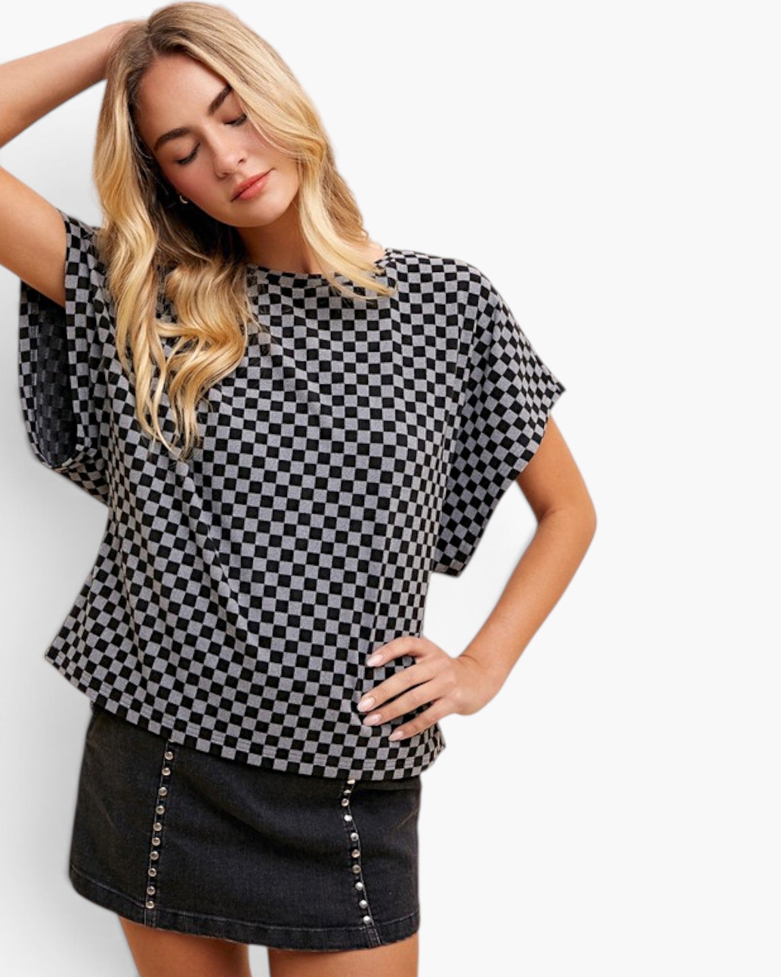 A woman modeling a black and white checkerboard short-sleeve top with a relaxed fit, paired with a dark denim skirt, against a plain white studio background.