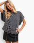 A woman modeling a black and white checkerboard short-sleeve top with a relaxed fit, paired with a dark denim skirt, against a plain white studio background.