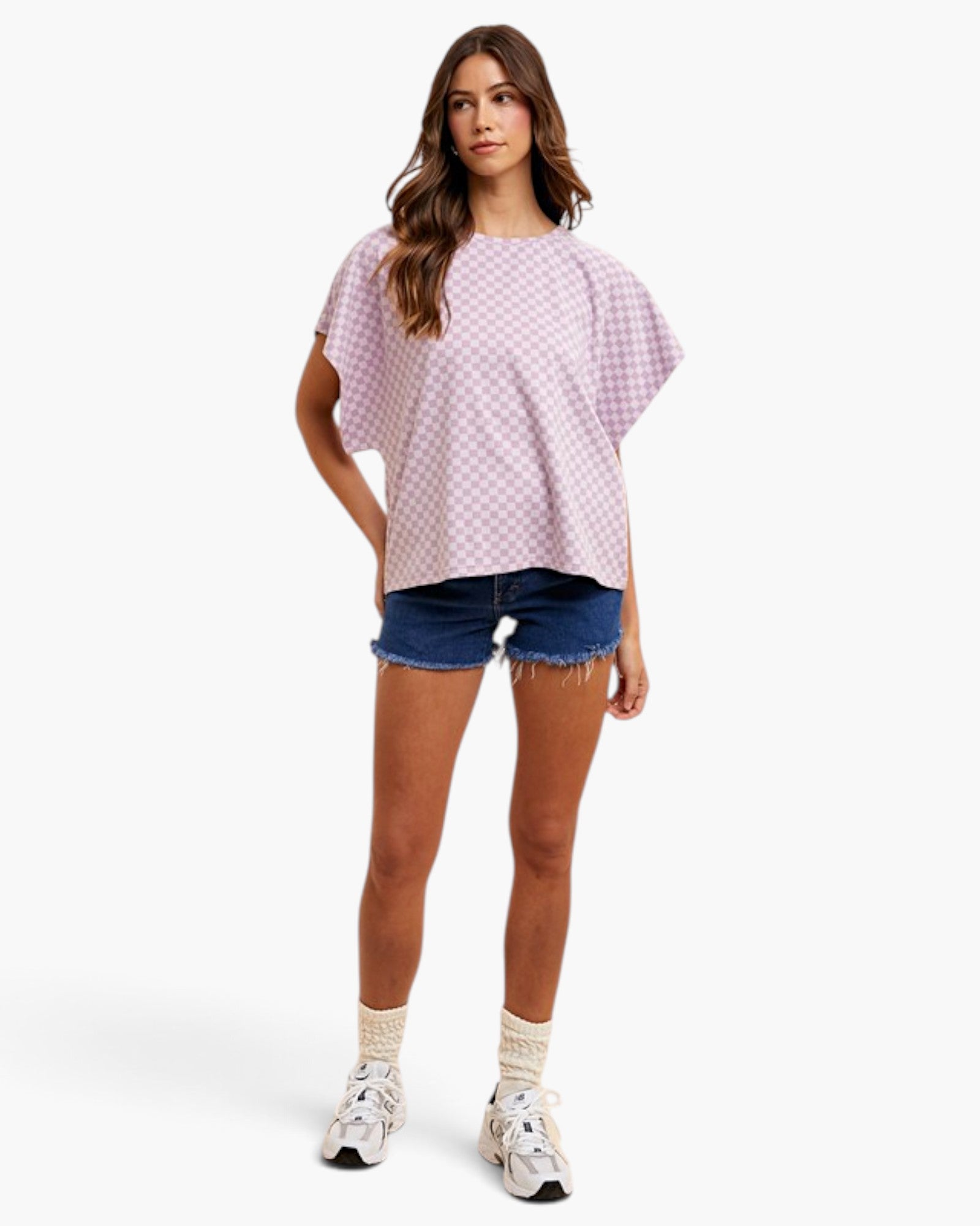 A woman modeling a short-sleeve, loose-fitting top with a pink and white gingham pattern, paired with denim shorts, white socks, and white sneakers, against a plain white studio background.
