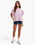 A woman modeling a short-sleeve, loose-fitting top with a pink and white gingham pattern, paired with denim shorts, white socks, and white sneakers, against a plain white studio background.
