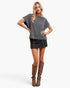 A woman modeling a black and white checkered short-sleeve top with a black denim mini skirt and brown knee-high boots, posed against a light gray studio background.