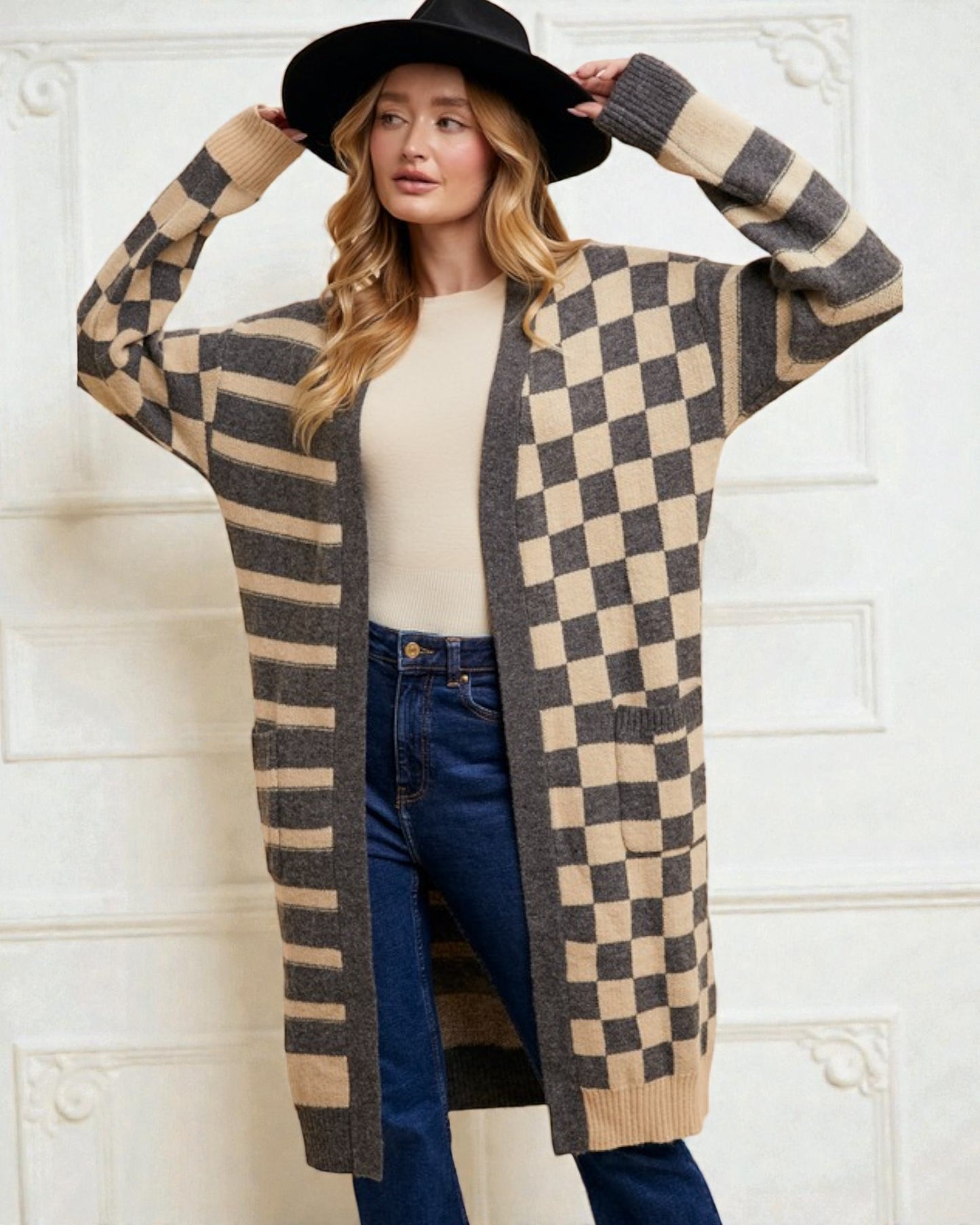 Woman wearing a checkered cardigan, black hat, and blue jeans against a white wall.
