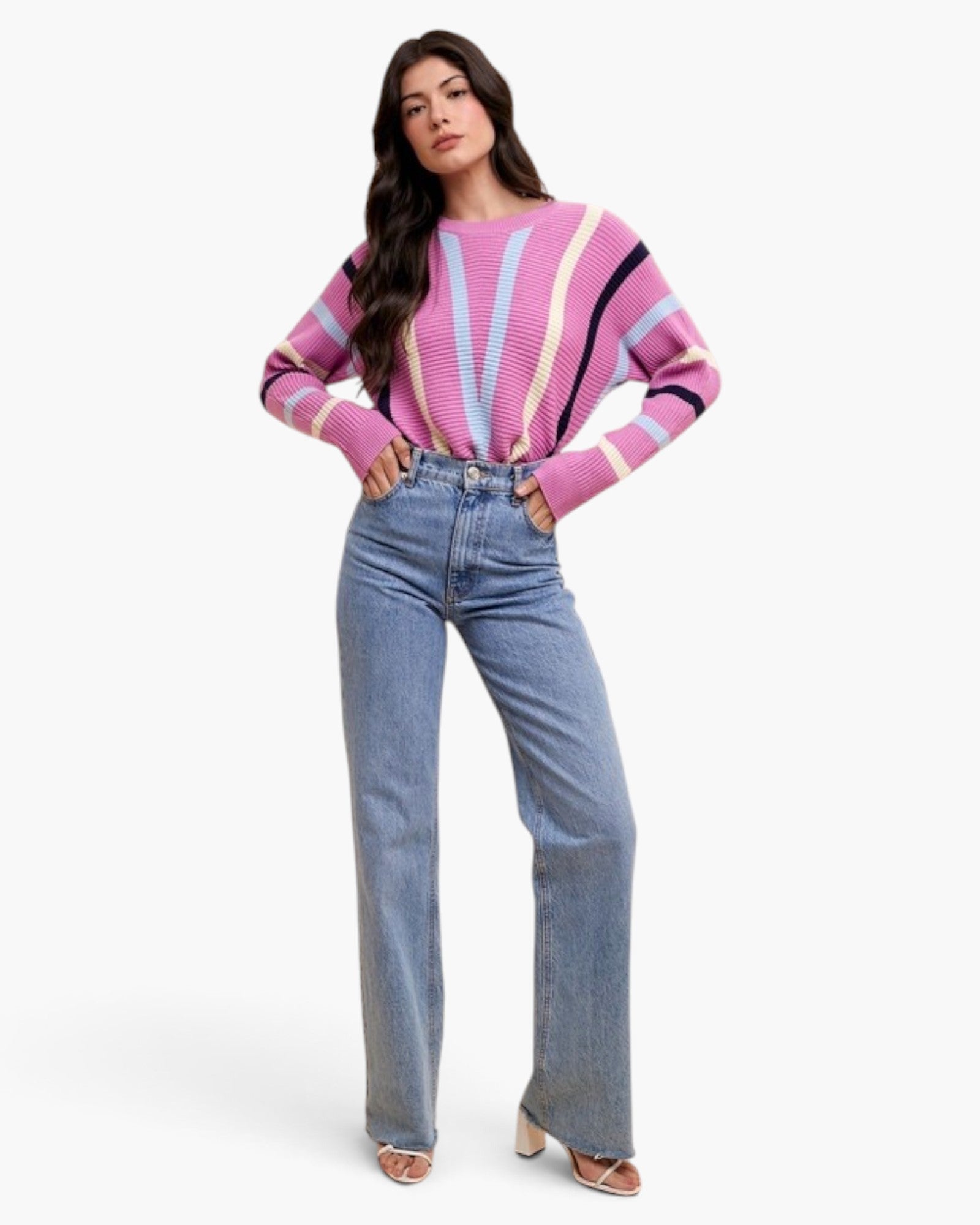 A woman modeling a pink striped knit sweater with blue jeans and beige heeled sandals, standing against a plain light gray studio background.