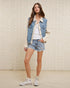 Woman wearing a denim jacket, white shirt, and shorts against a light brick wall.