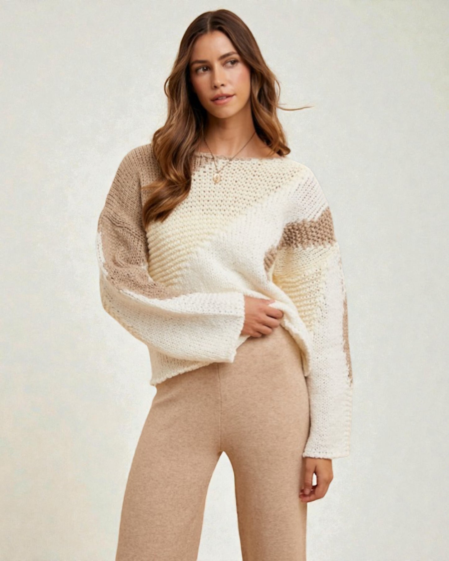 Woman wearing a beige and white sweater with beige pants on a plain background