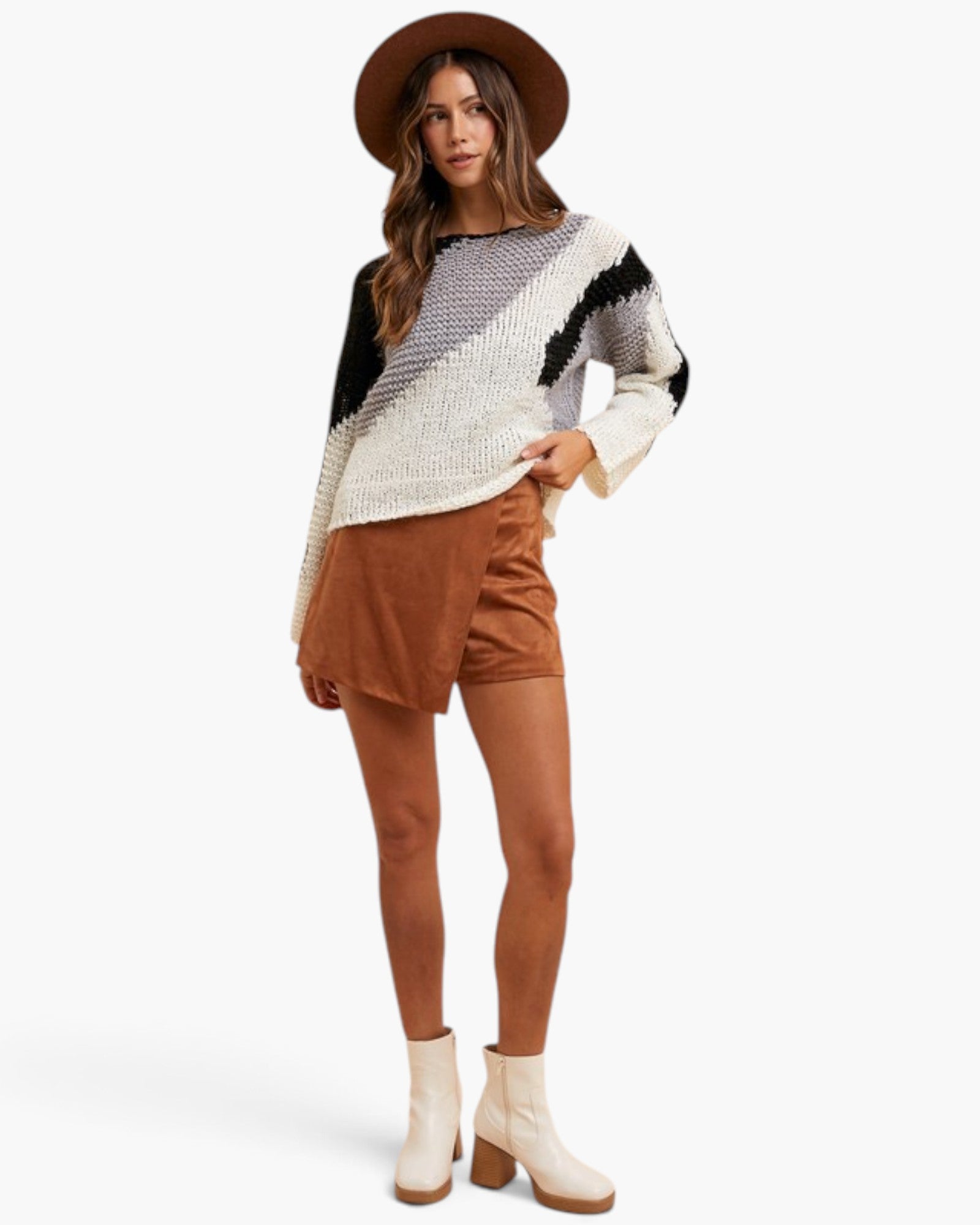 A woman modeling a chunky knit sweater with black, white, and gray geometric patterns, paired with brown shorts, a brown wide-brim hat, and white ankle boots, set against a plain white studio background.