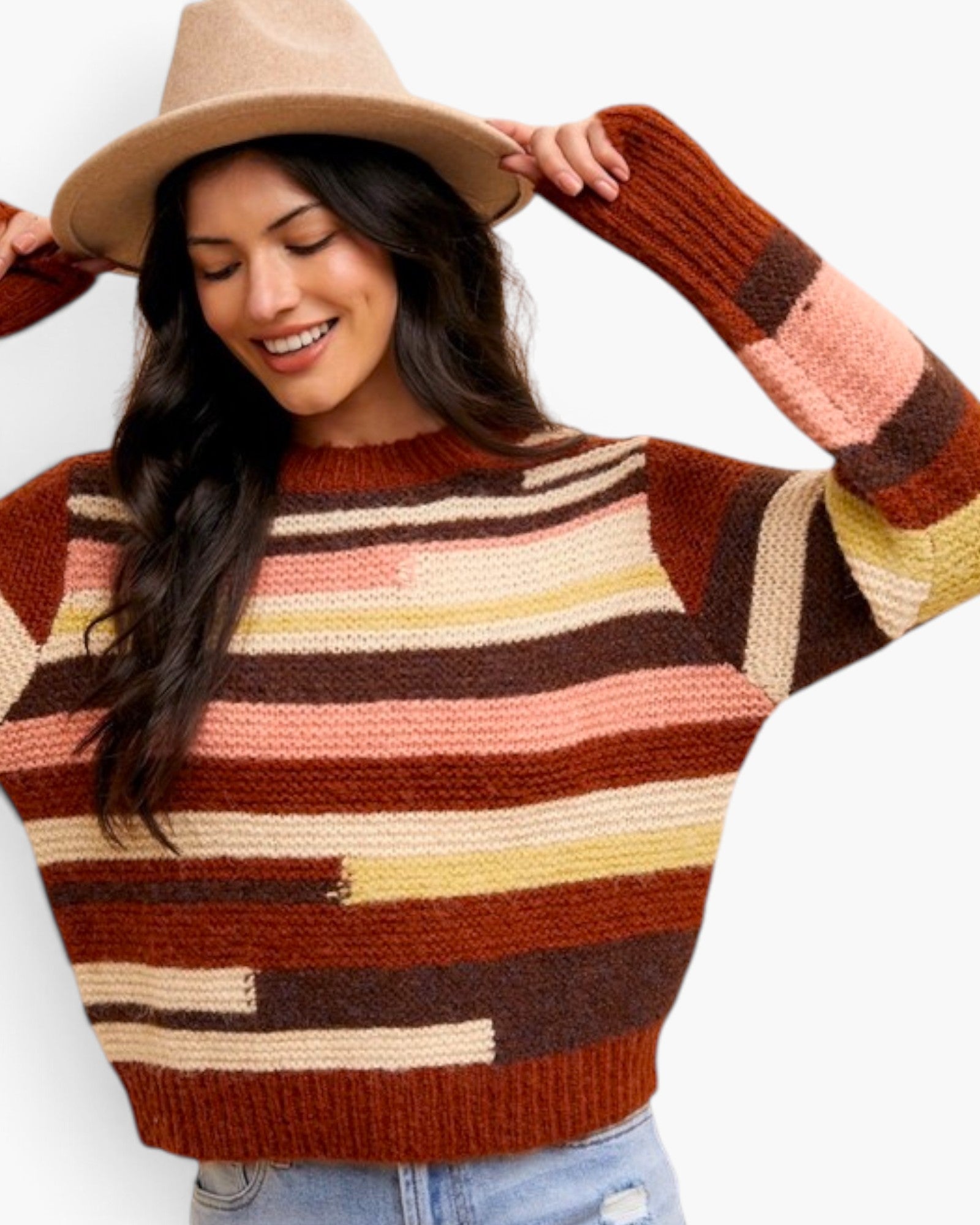 A woman wearing a chunky knit sweater with horizontal stripes in rust brown, dark brown, beige, and soft pink, paired with a tan wide-brim hat and light blue jeans against a plain white background.