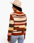 A woman shown from the back wearing a rust-brown knit sweater with horizontal stripes in cream, pink, mustard yellow, and dark brown, paired with light-wash blue jeans and a beige wide-brim hat against a white studio background.