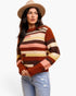 A woman wearing a chunky knit sweater with horizontal stripes in rust orange, brown, cream, and dusty pink, paired with light blue jeans and a tan wide-brim hat, against a plain white background.