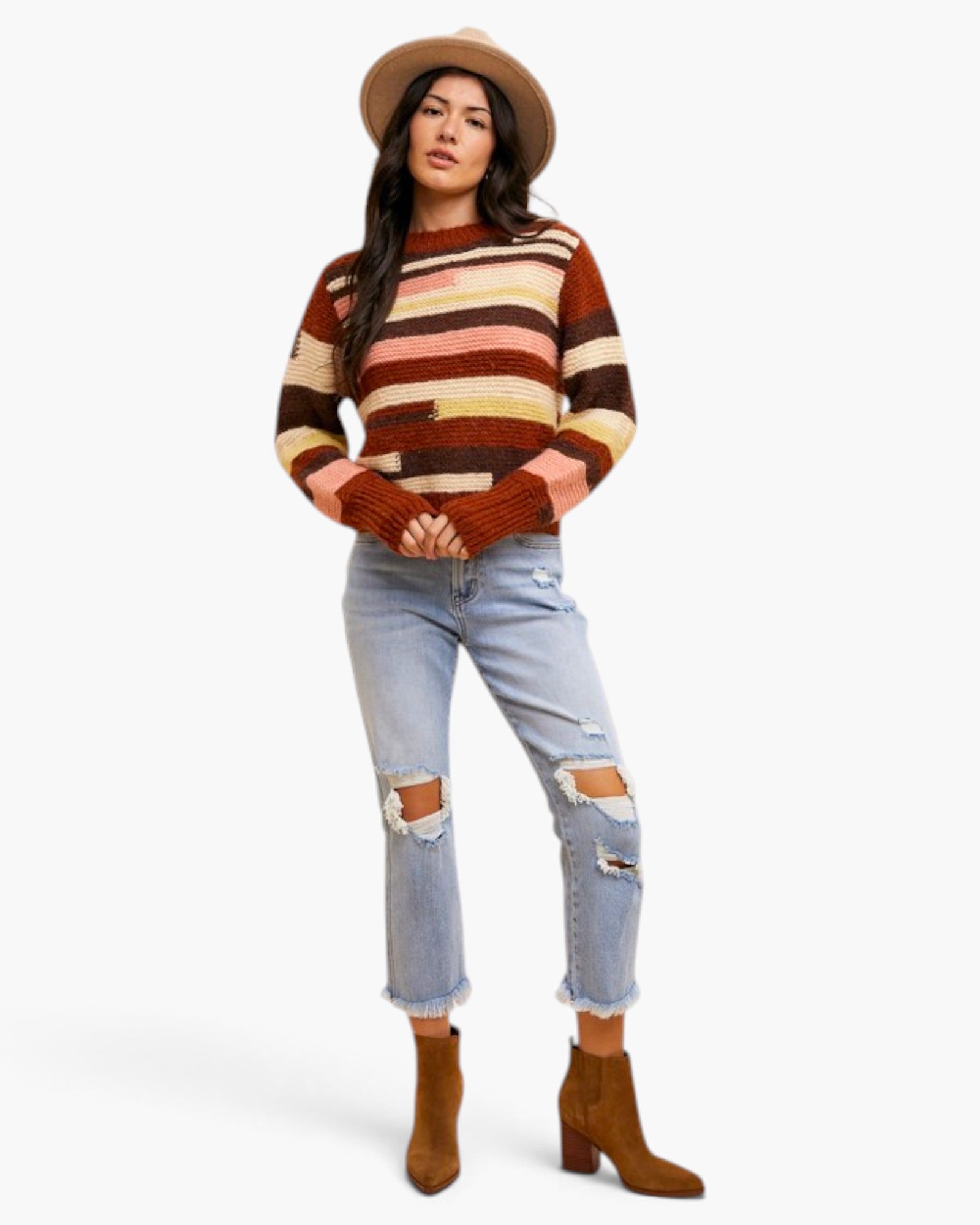 A woman modeling a striped knit sweater with horizontal bands of rust brown, tan, cream, and dusty pink, paired with light-wash distressed jeans, brown ankle boots, and a tan wide-brim hat against a plain white studio background.