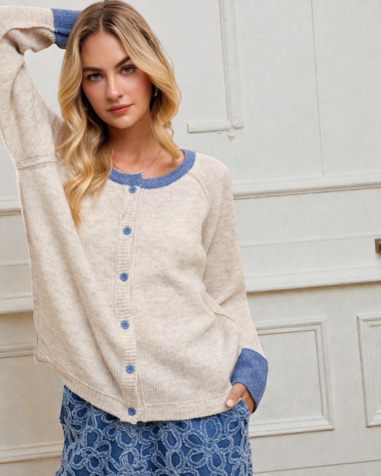 Woman wearing a cream cardigan with blue buttons and trim, standing against a white wall.