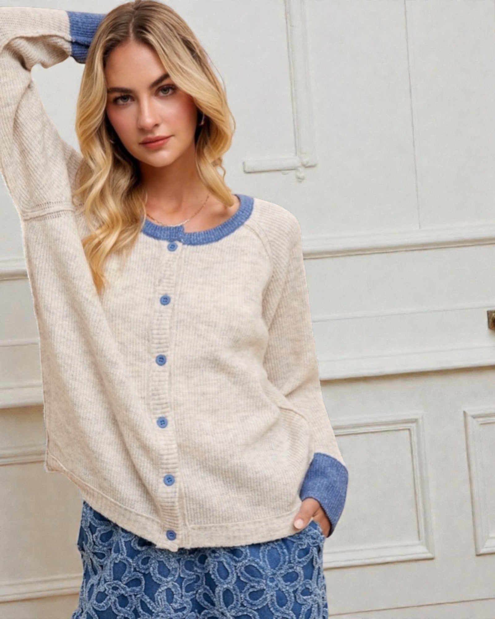 Woman wearing a cream cardigan with blue buttons and trim, standing against a white wall.