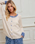 Woman wearing a cream cardigan with blue buttons and trim, standing against a white wall.
