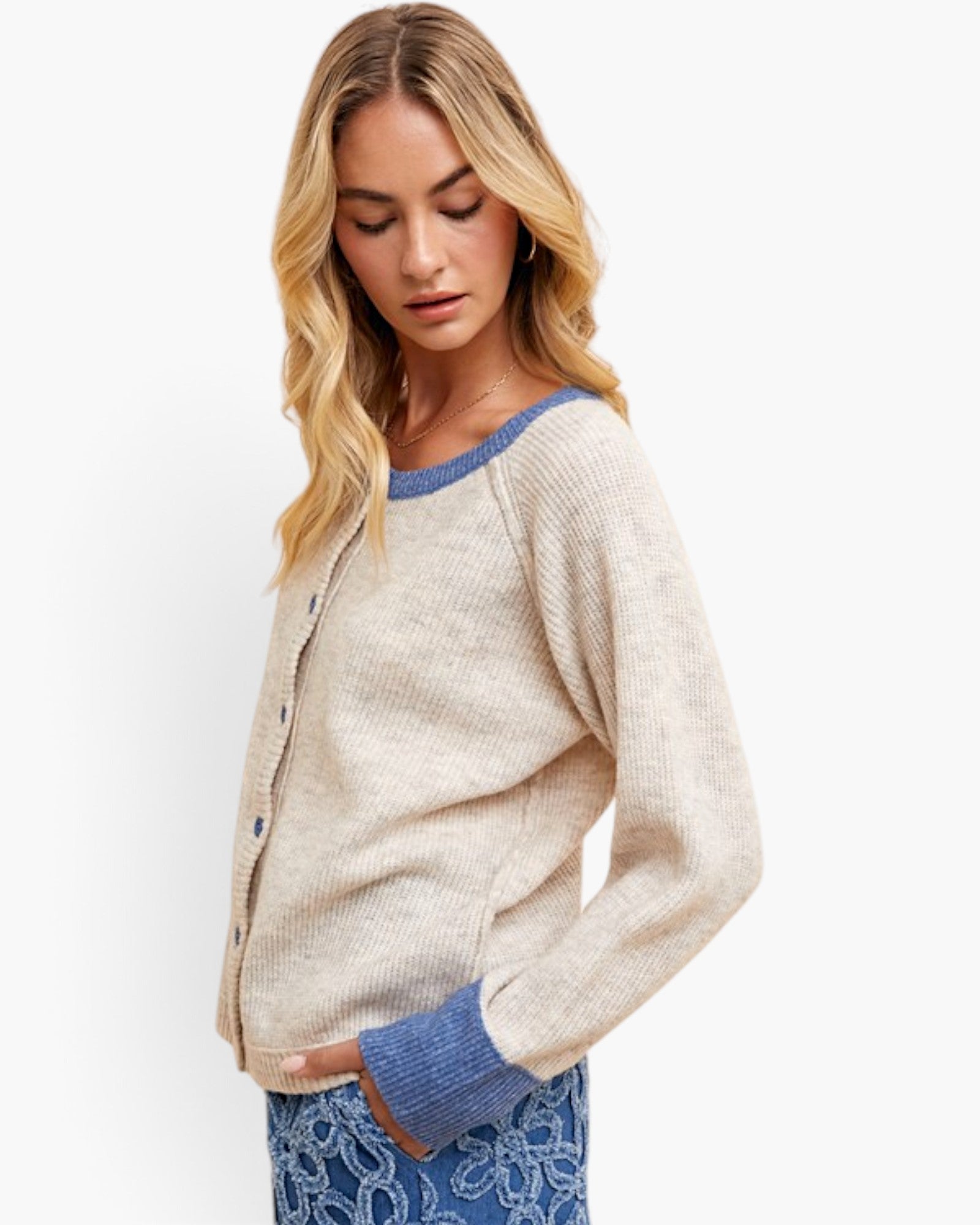 A woman wearing a beige knit cardigan with blue ribbed trim and cuffs, paired with blue patterned pants, against a plain white studio background.