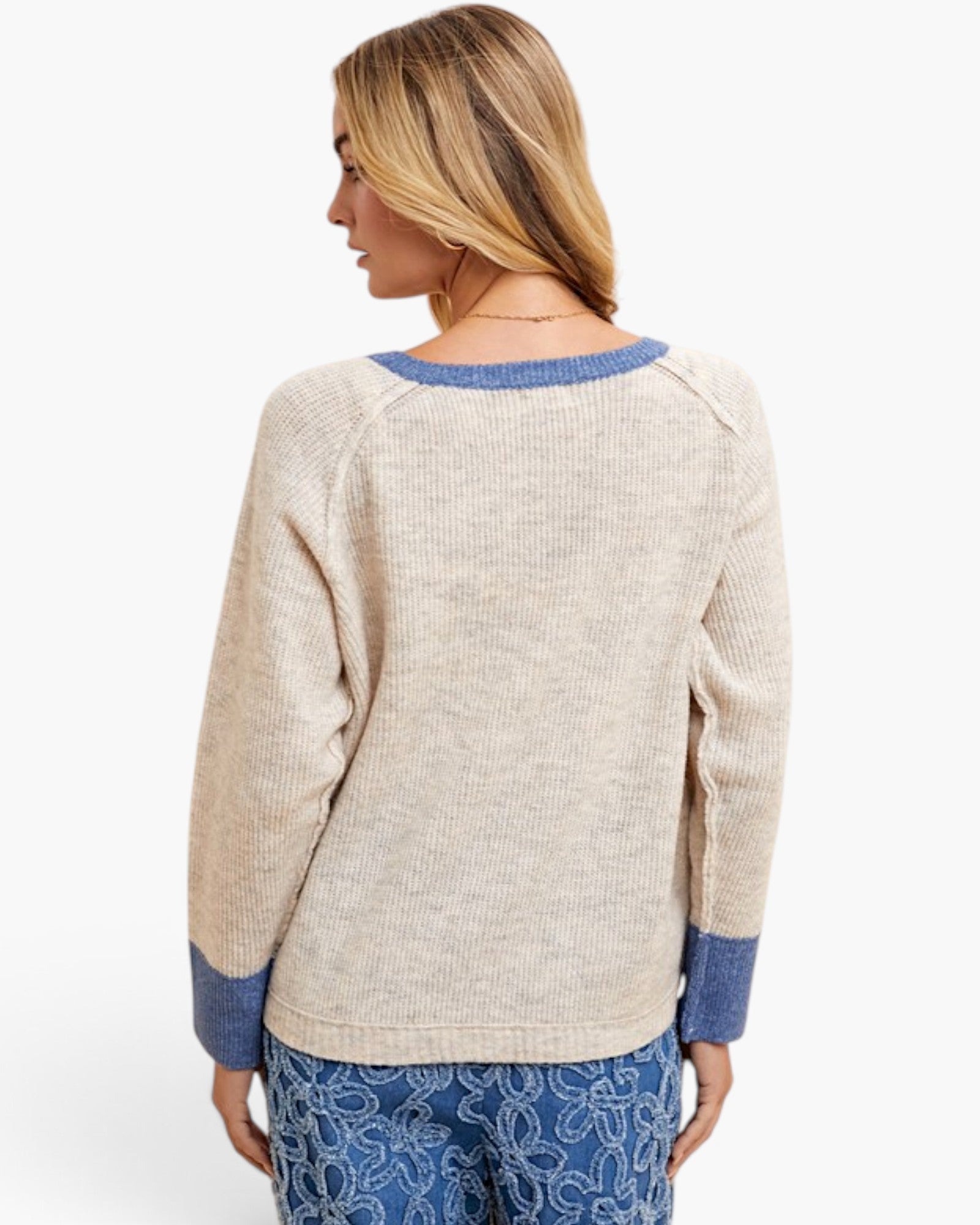 A woman shown from the back wearing a beige knit sweater with blue ribbed trim at the crew neck, cuffs, and hem, paired with blue patterned pants against a light gray studio background.