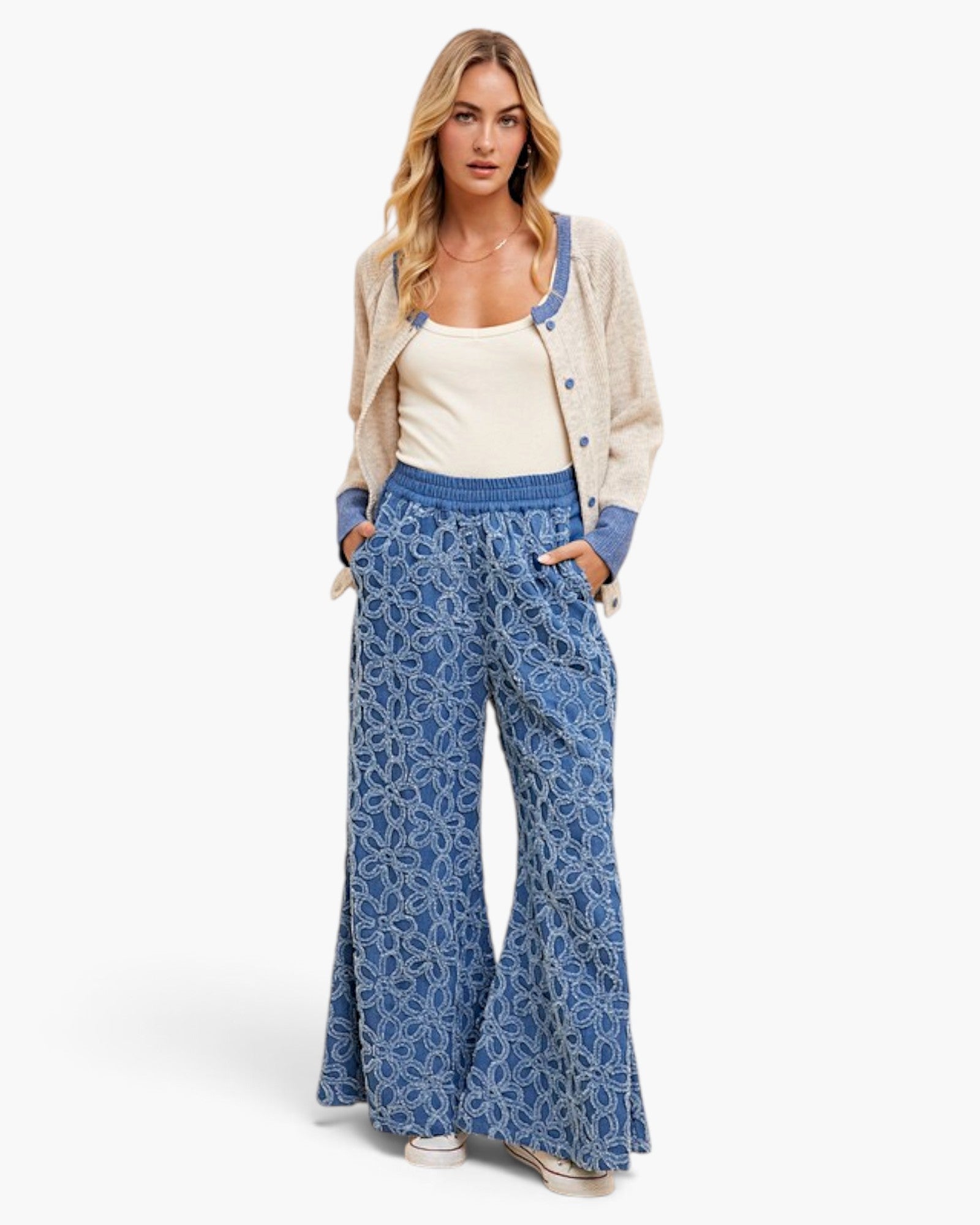 A woman modeling a casual outfit consisting of wide-leg blue pants with a white floral pattern, a cream tank top, a beige cardigan with blue trim, and white sneakers, set against a plain white studio background.