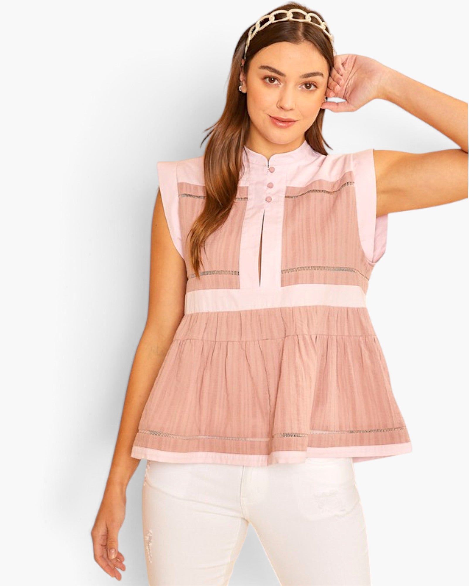 A woman modeling a sleeveless pink peplum top with a mock neck, button placket, and horizontal striped details, paired with white pants against a light gray studio background.