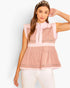 A woman modeling a sleeveless pink peplum top with a mock neck, button placket, and horizontal striped details, paired with white pants against a light gray studio background.