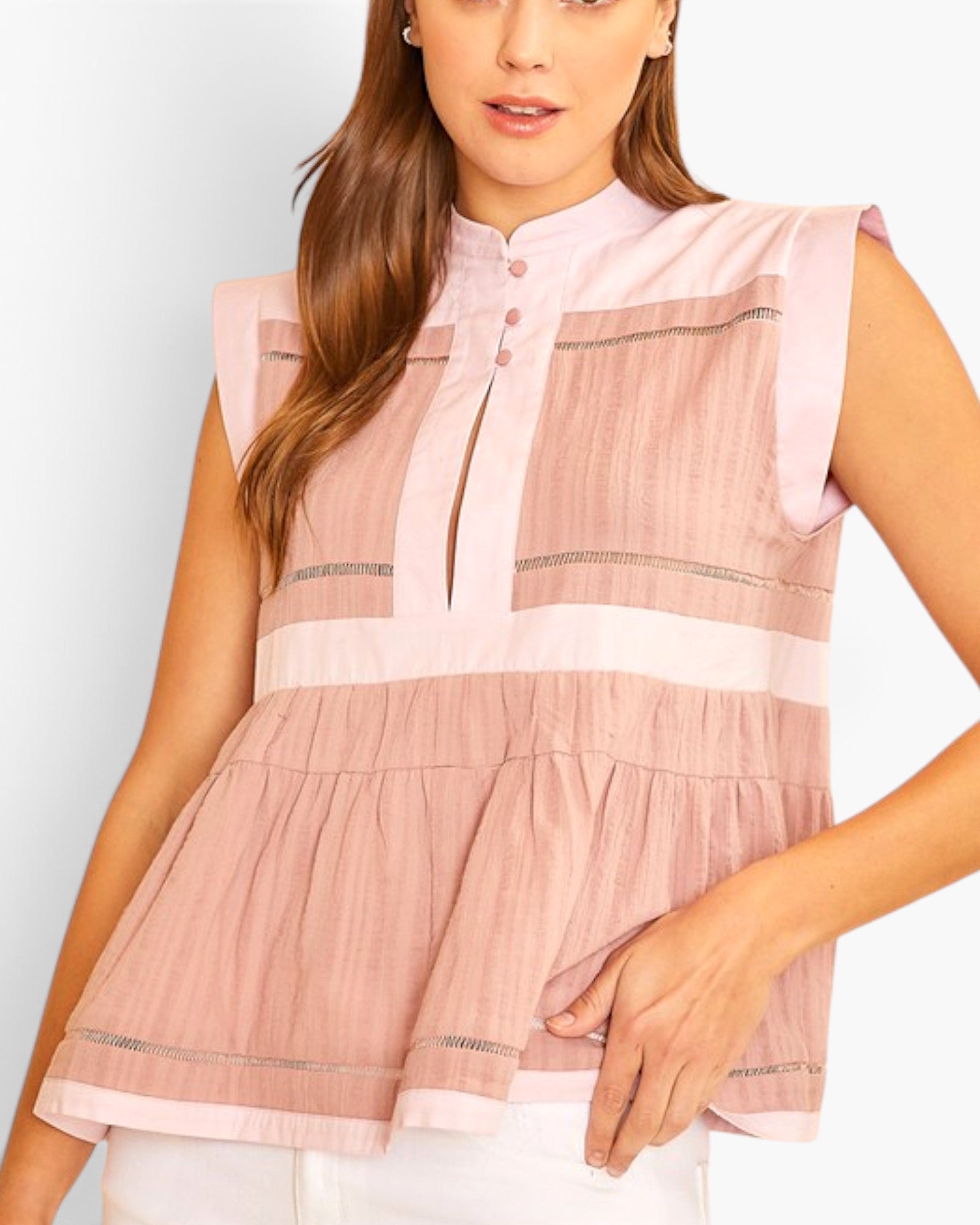 A woman wearing a sleeveless pink peplum top with a mock neck, button placket, and horizontal textured stripe panels, paired with white pants against a light gray studio background.