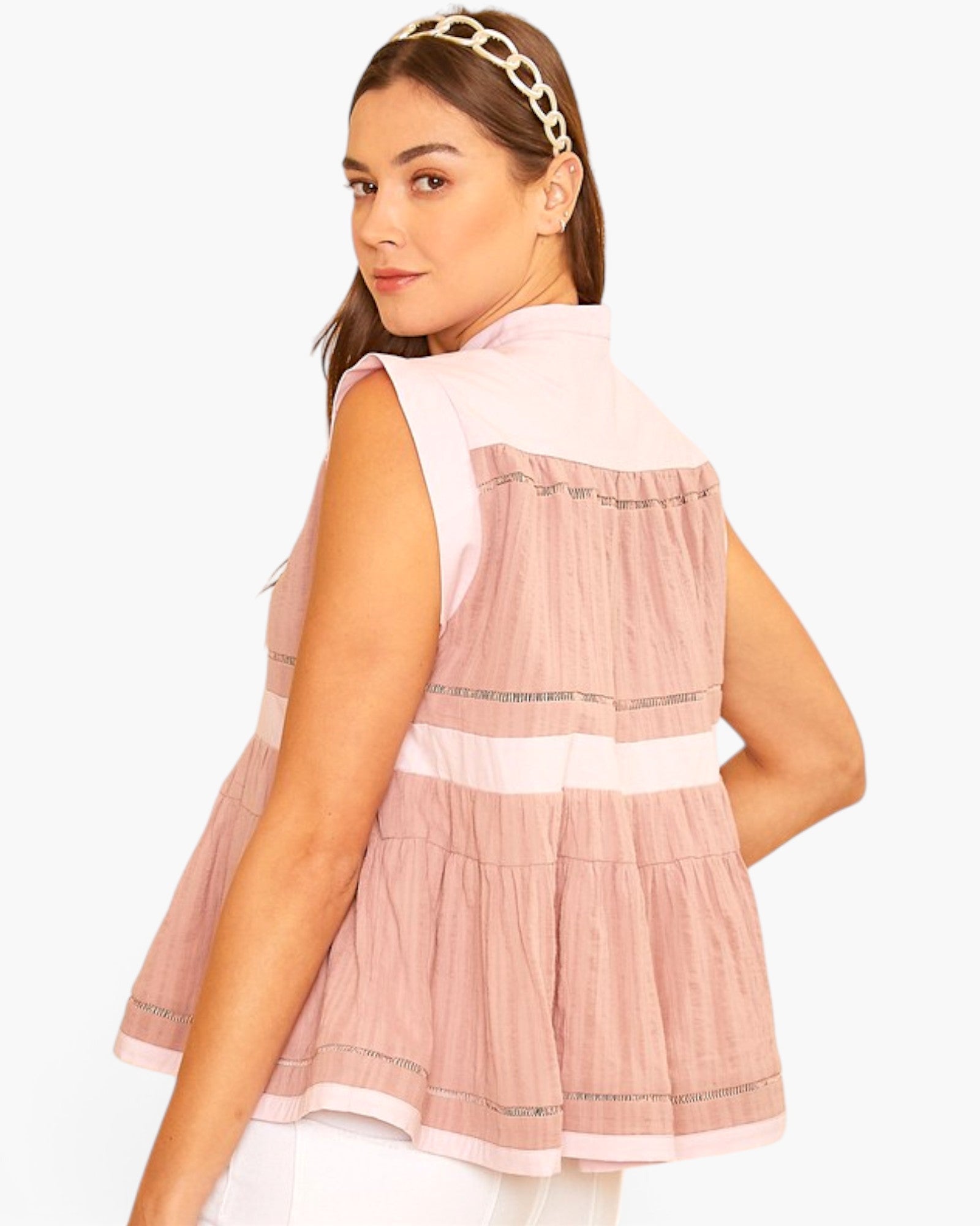 A woman wearing a sleeveless tiered peplum top in light pink and white with thin metallic trim, shown from a three-quarter back view against a plain white background.
