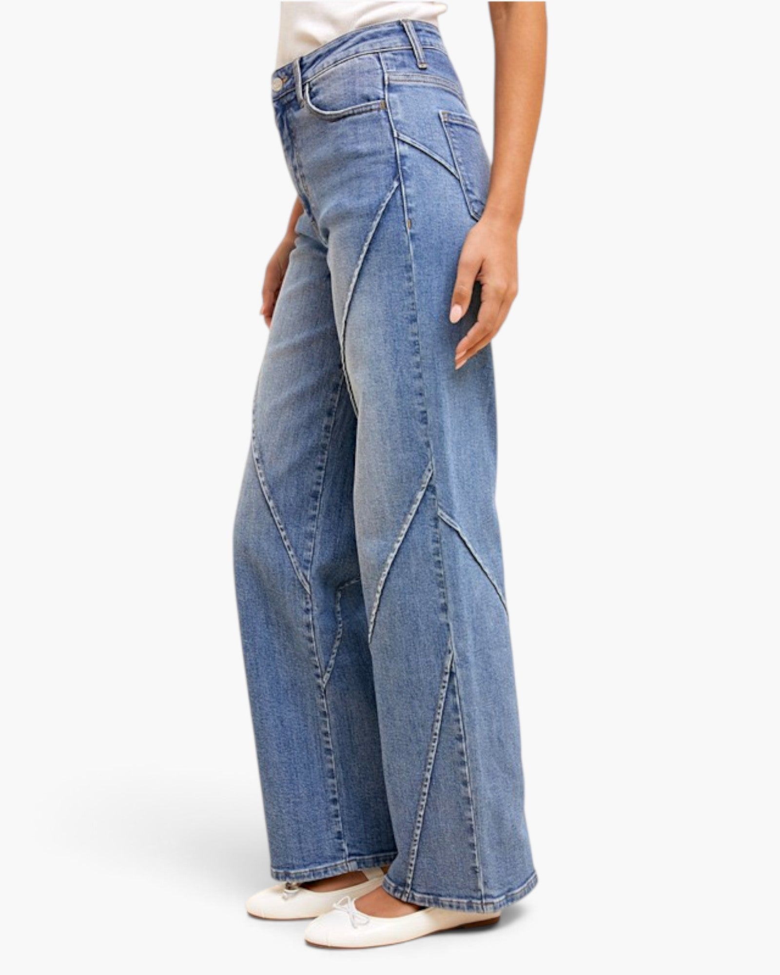 A person wearing light blue wide-leg denim jeans with diagonal seam detailing, paired with white flats, shown against a plain light gray studio background.