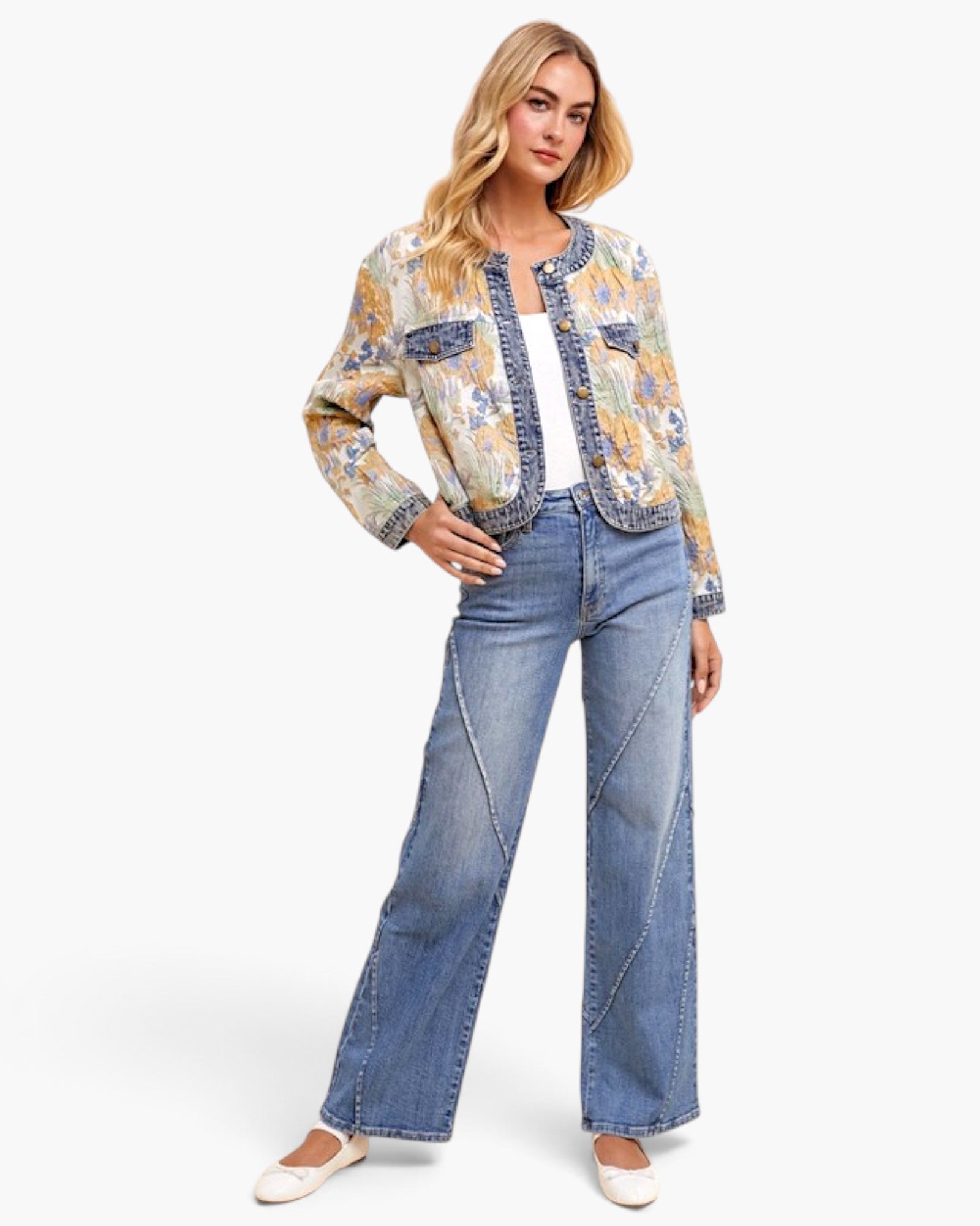 A woman modeling a cropped, long-sleeve denim jacket with a colorful abstract print in yellow, blue, and orange, paired with wide-leg blue jeans and white flats, against a light gray studio background.