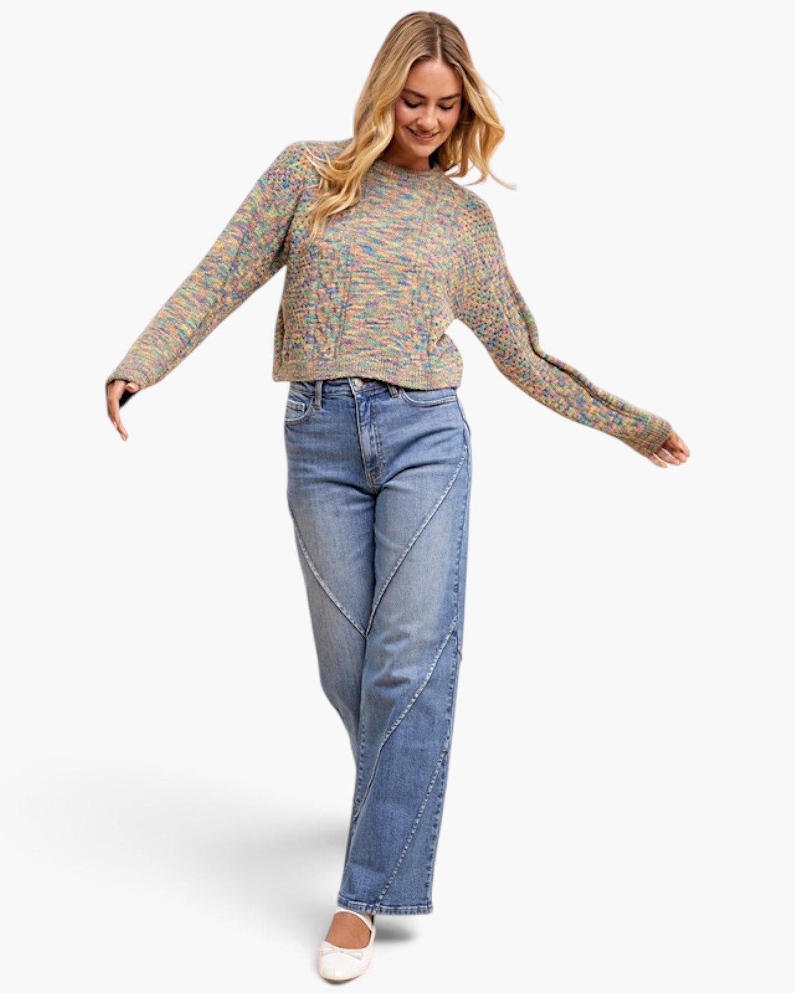 A woman modeling a long-sleeve knit sweater with a multicolor abstract pattern in beige, green, pink, and blue, paired with light-wash blue jeans and white flats, against a plain white studio background.