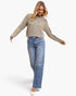 A woman modeling a long-sleeve knit sweater with a multicolor abstract pattern in beige, green, pink, and blue, paired with light-wash blue jeans and white flats, against a plain white studio background.