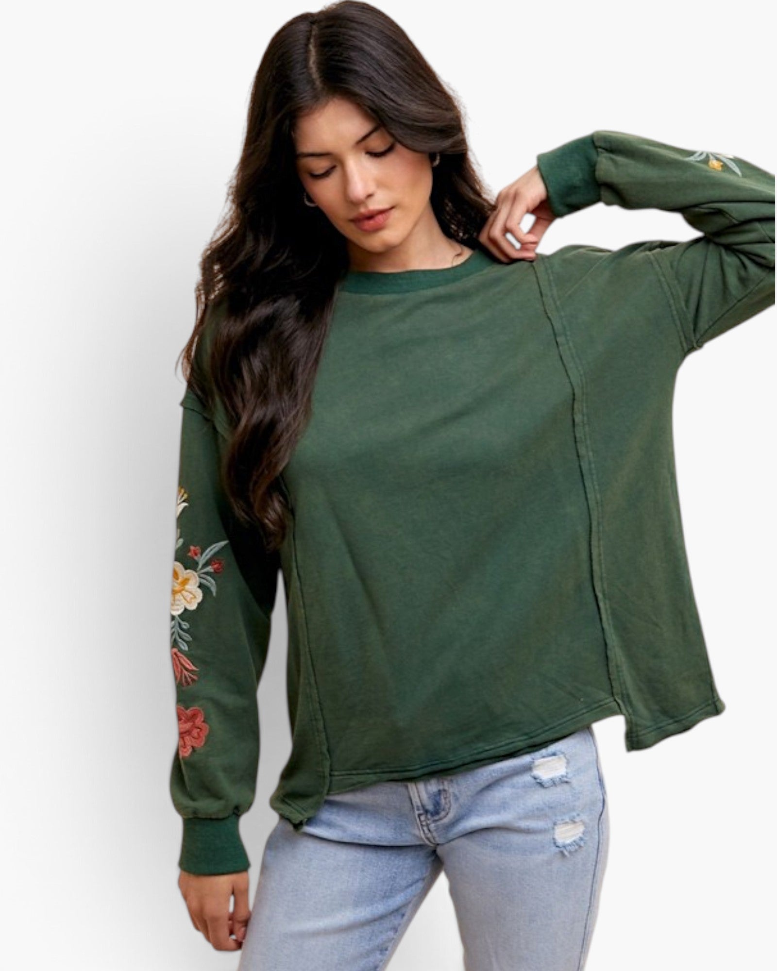 A woman wearing a dark green long-sleeve top with floral embroidery on one sleeve, paired with light blue jeans, against a plain white background.