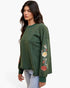 A woman wearing an olive green long-sleeve sweatshirt with colorful floral embroidery on the sleeve, paired with light blue jeans, photographed against a plain white background.